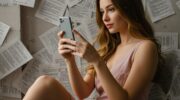 Женщина сидит с телефоном в руках, задумчиво смотрит в камеру, вокруг кружатся листы с недописанными сообщениями
